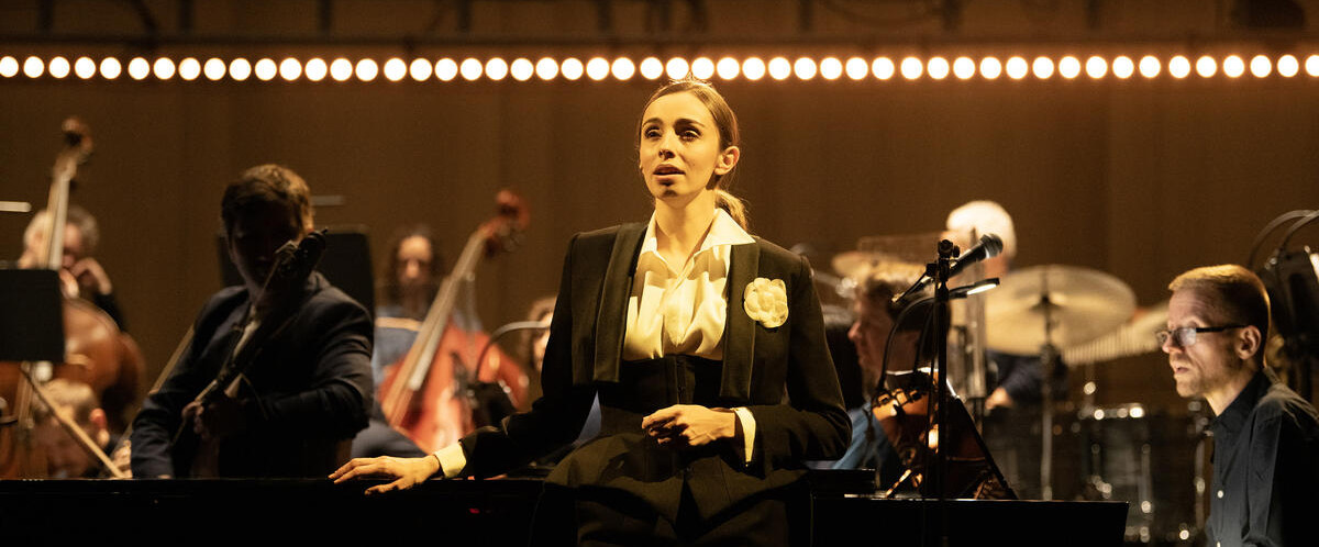 Lea Desandre est adossée au piano, en tenue de queue de pie. Derrière elle, on aperçoit une partie de l'orchestre